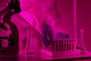 Scientist in a lab coat handling test tubes under pink lighting, using a microscope.