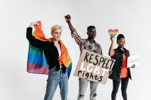 Three adults holding signs and a pride flag promoting LGBT rights on a white background.